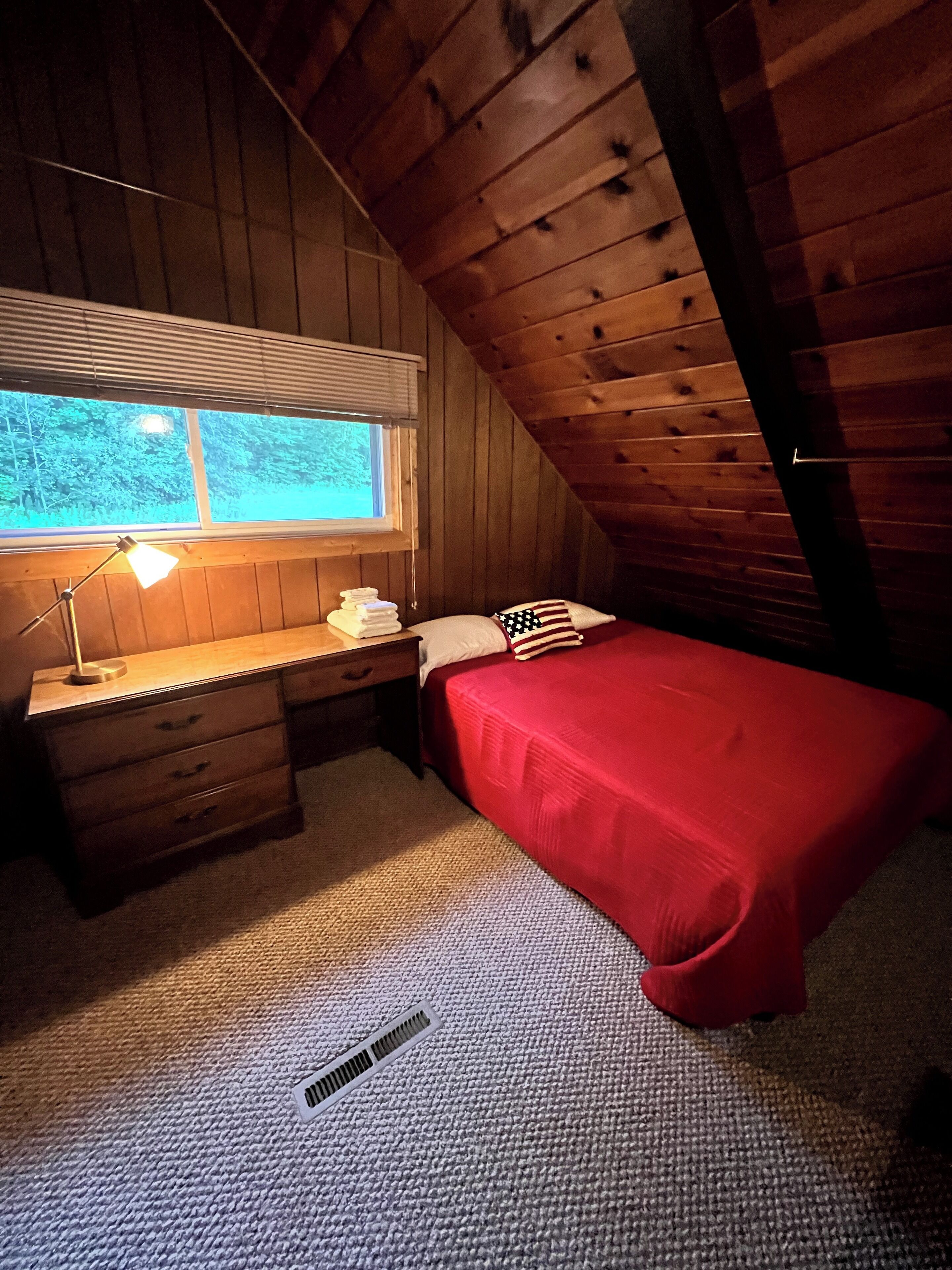 A small, cozy attic bedroom with a wooden sloped ceiling, a single bed with a red bedspread, a desk with a lamp, and a window with blinds.