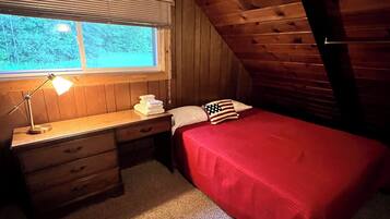 A small, cozy attic bedroom with a wooden sloped ceiling, a single bed with a red bedspread, a desk with a lamp, and a window with blinds.