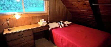 Un dormitorio acogedor en el ático, con techo inclinado de madera, una cama individual con colcha roja, un escritorio con lámpara y una ventana con persianas.