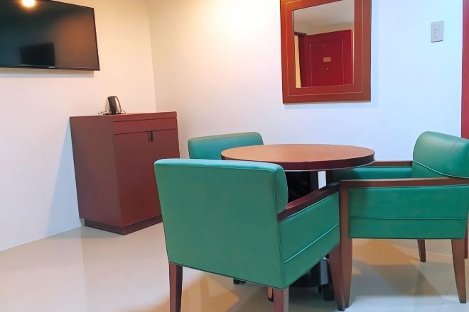 Um quarto com uma mesa de madeira redonda, duas cadeiras verdes, um armário vermelho e uma TV montada na parede.