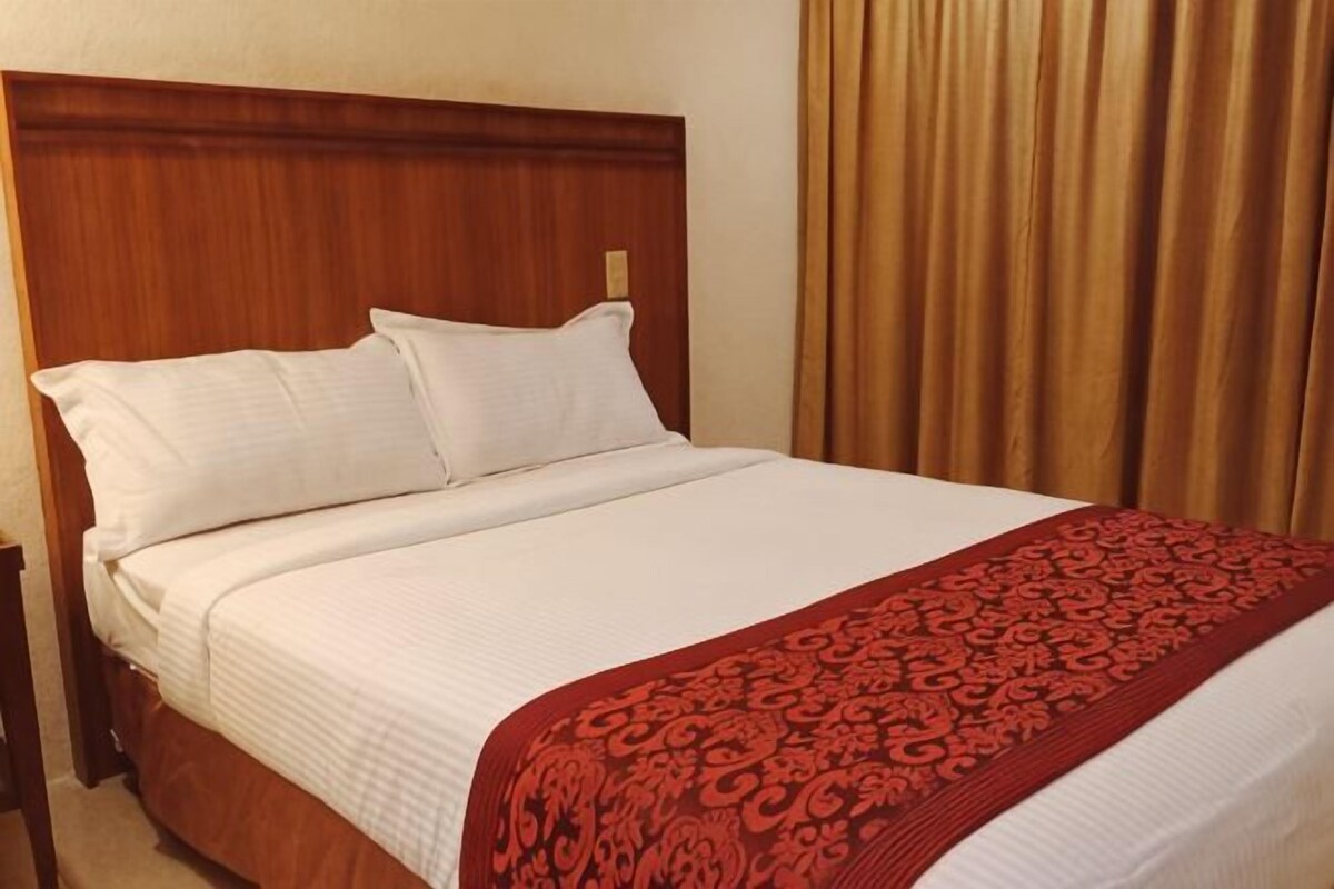 A hotel room with a wooden headboard, a large bed with white bedding, and a red patterned throw blanket.