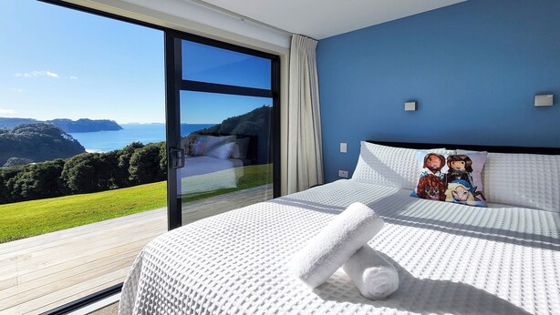 Un dormitorio con un ventanal grande, una cama con ropa de cama blanca y vistas al mar y las colinas.