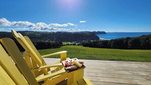 Una silla Adirondack amarilla sobre una terraza de madera con vistas a un paisaje costero, cielo despejado y sol brillante.
