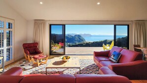 Amplia sala de estar con un sofá rojo, una mesa de centro de madera y una alfombra colorida. Un amplio ventanal corredero da a un área exterior con vistas al mar y colinas.