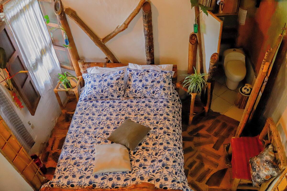 Una habitación rústica con una cama, una silla de madera, una mesita pequeña y un inodoro.