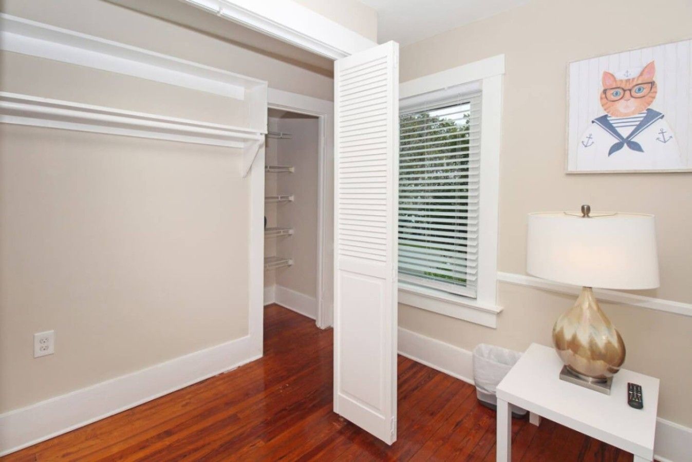 A room with a wooden floor, a white door, a window with blinds, a lamp on a table, and a painting on the wall.