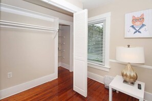 Una habitación con piso de madera, una puerta blanca, una ventana con persianas, una lámpara sobre una mesa y un cuadro en la pared.