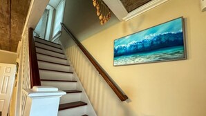 Una escalera con escalones de madera blancos y oscuros, barandal blanco y un cuadro anclado a la pared que representa una escena marina azul.