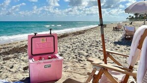 Escena de playa con un enfriador rosa, una sombrilla a rayas y una silla de playa de madera.