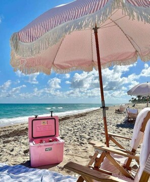 Eine Strandansicht mit einer pinken Kühlbox, einem gestreiften Strandsonnenschirm und einem hölzernen Liegestuhl.