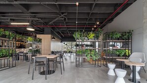 Ein modernes Café mit mehreren Sitzbereichen, darunter Tische und Stühle, umgeben von Regalen mit Pflanzen.
