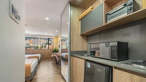 Eine kompakte Küchenzeile mit Mikrowelle, kleinem Kühlschrank und Spüle. Es gibt einen Essbereich mit Tisch und Stühlen sowie einen Blick auf die Stadtlandschaft durch ein großes Fenster.