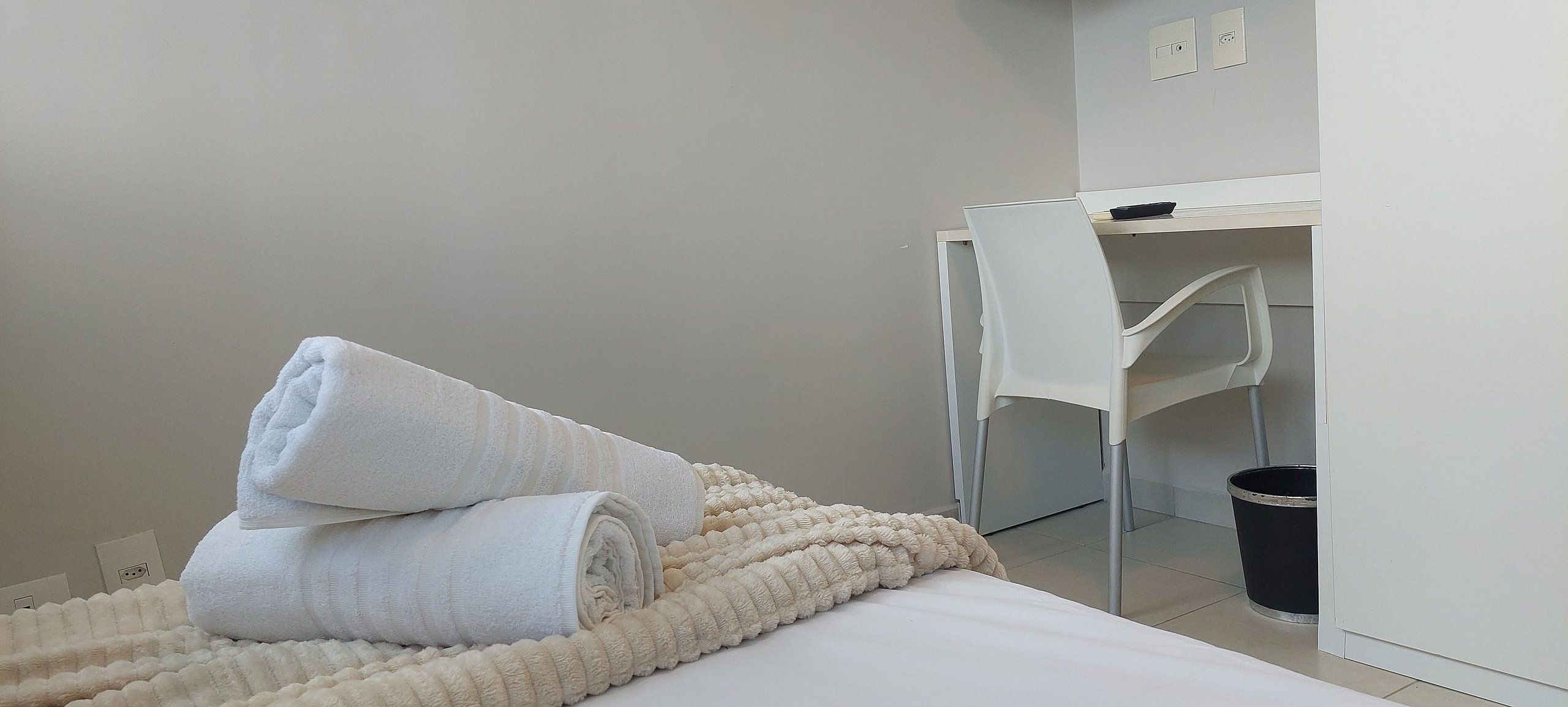 Une chambre d’hôtel avec un lit, une chaise blanche, un bureau et une poubelle.