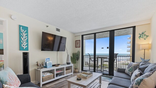 Ein Wohnzimmer mit großem Fernseher, Couchtisch und Sofa. Ein Balkon bietet Blick auf den Strand.