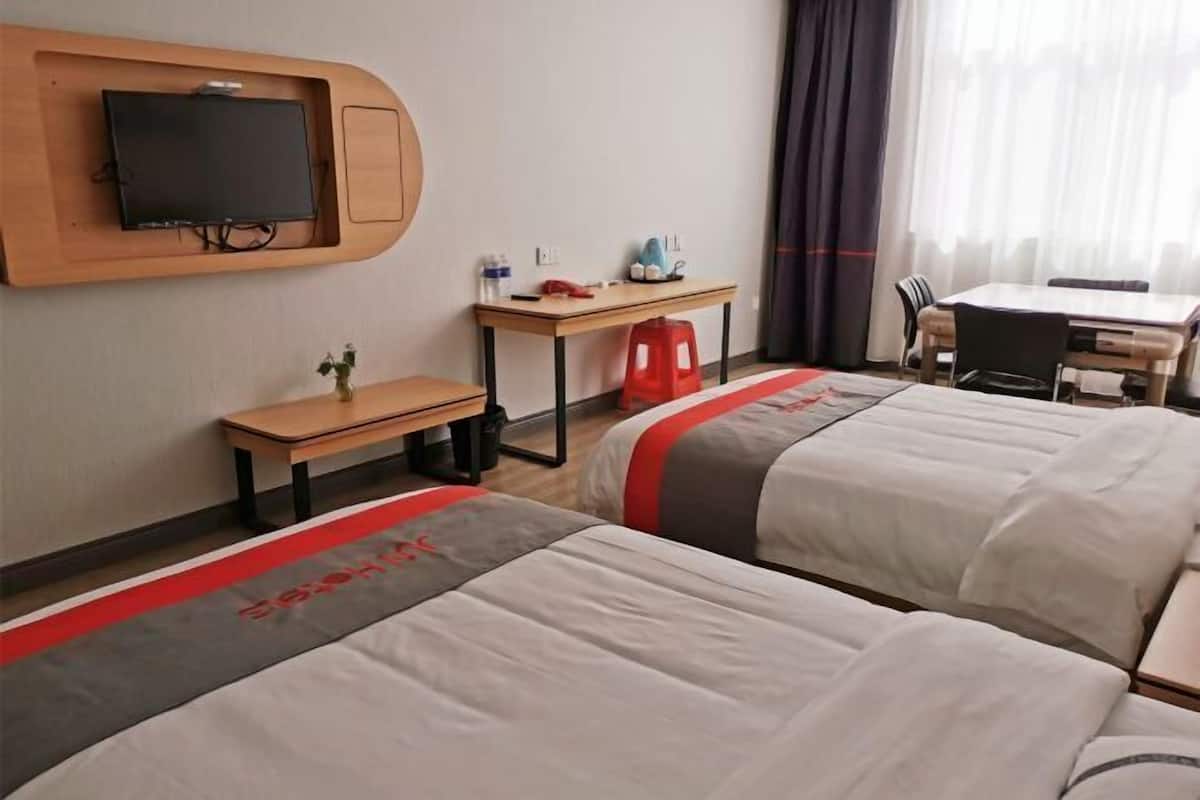 A hotel room with two beds, a TV mounted on the wall, a small desk with a chair, and a dining table with chairs.