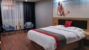 A hotel room with a large bed, two blue chairs, a small table, and a painting of a beach scene on the wall. - Junyi Chain Hotel (Tianmen baoan palza Store) (Tianmen)