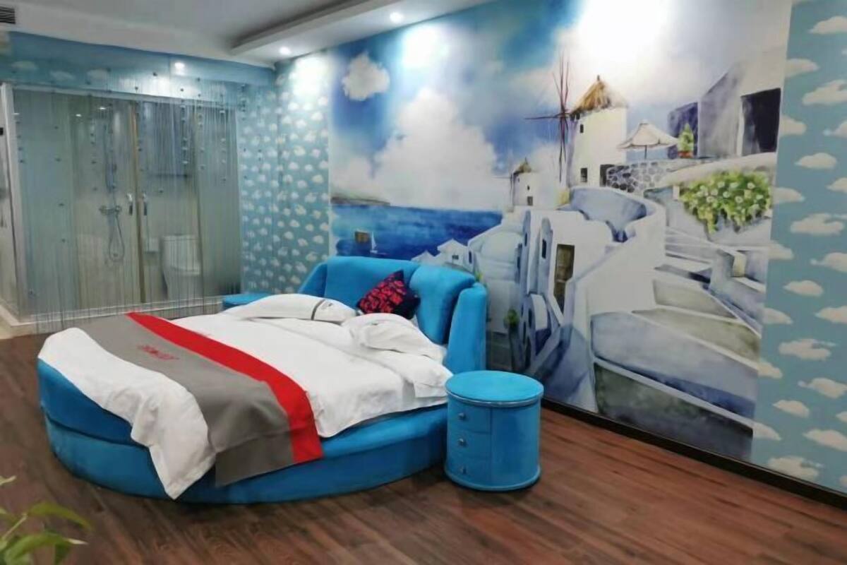 A bedroom with a bed, a blue chair, a blue side table, and a glass-enclosed shower.