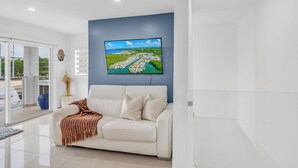 A modern living room with a white sofa, a television mounted on a blue wall, and a view of a green landscape through a glass door. - *NEW* Casa Coco-Pool/Tennis/Ramp (Key Largo)
