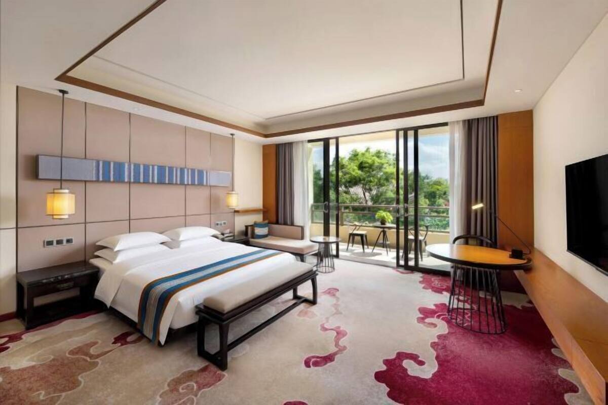 Ein Hotelzimmer mit Bett, Nachttischen, einem Schreibtisch, einem Sessel und einem Fernseher, sowie einem Balkon mit Blick.