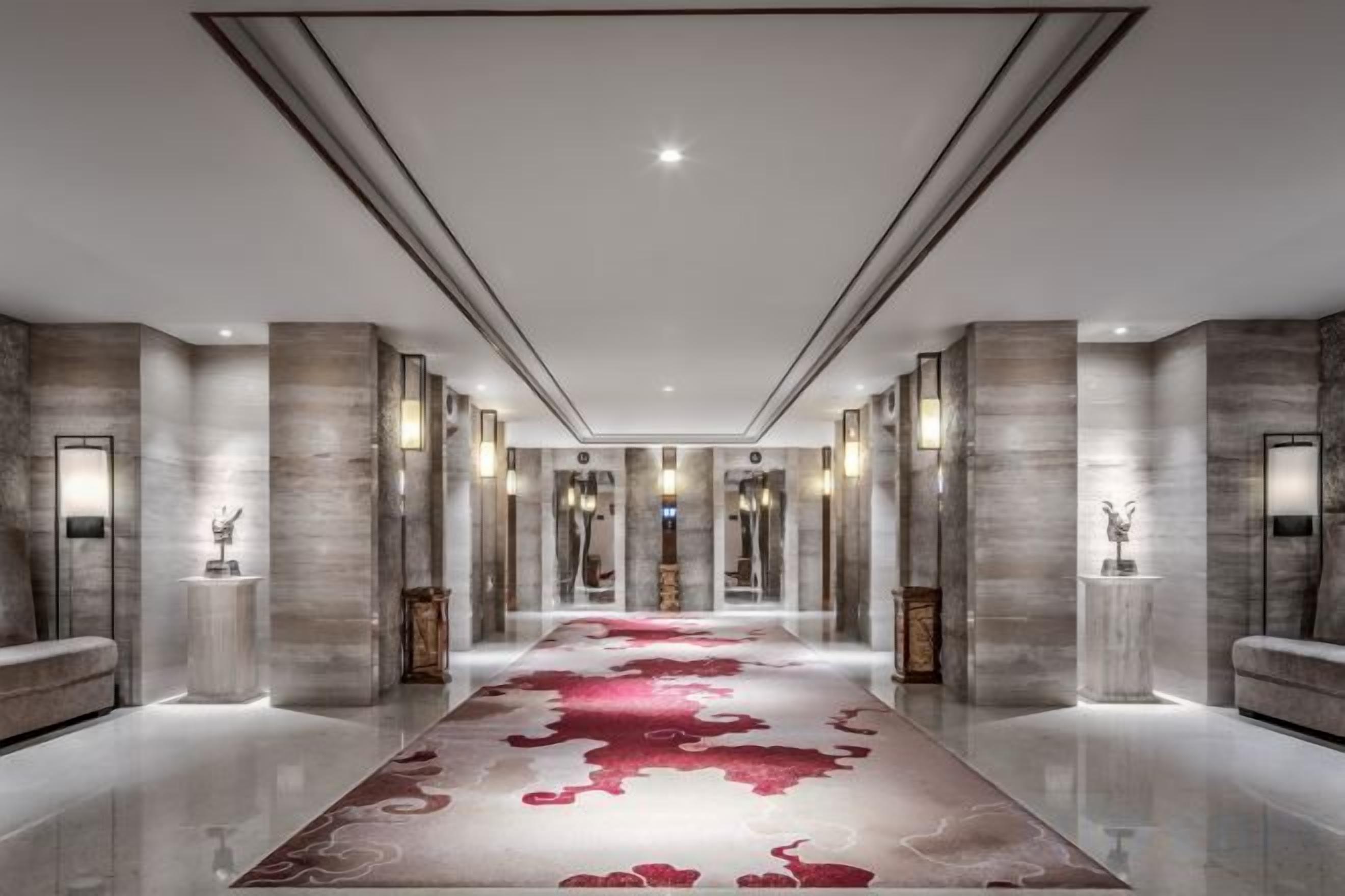 Un hall d’hôtel moderne avec un long couloir, des murs en marbre et une moquette rouge.