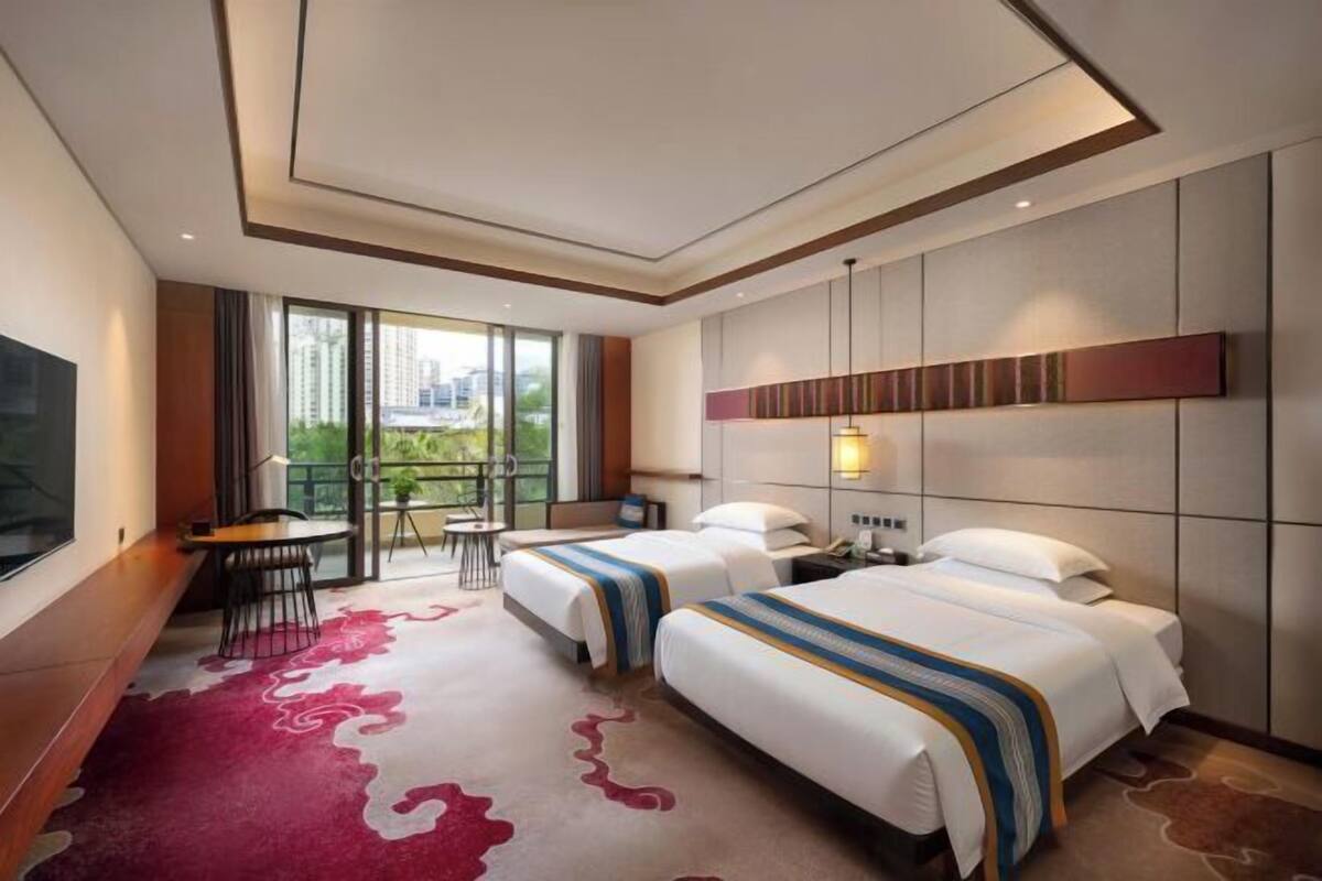 Ein Hotelzimmer mit zwei Betten, einem Fernseher, einem Schreibtisch und einem Balkon mit Blick auf die Stadt.