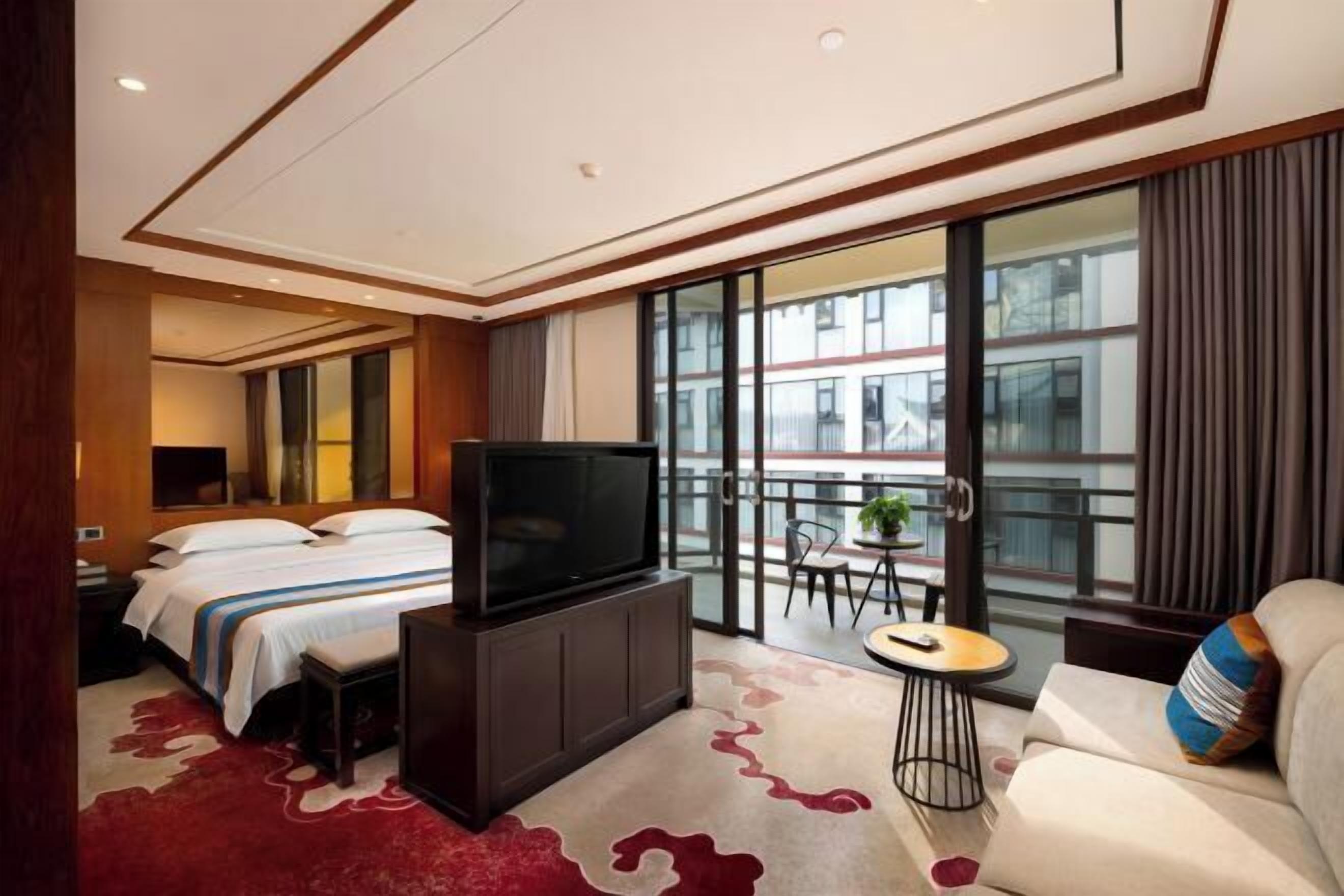 Een hotelkamer met een bed, een televisie, een bank en een balkon met uitzicht op gebouwen.