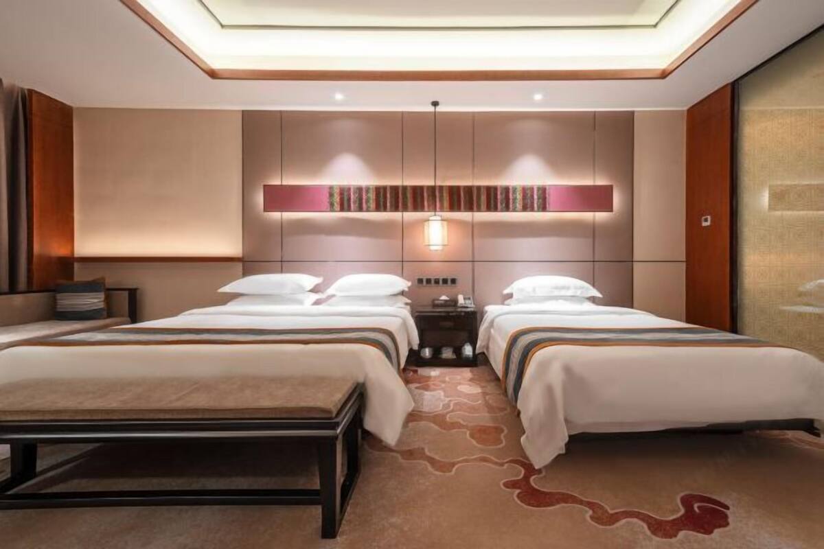 Ein Hotelzimmer mit zwei Betten, einer Sitzbank, einem Nachttisch, einem an der Wand befestigten Regal und einer Deckenleuchte.