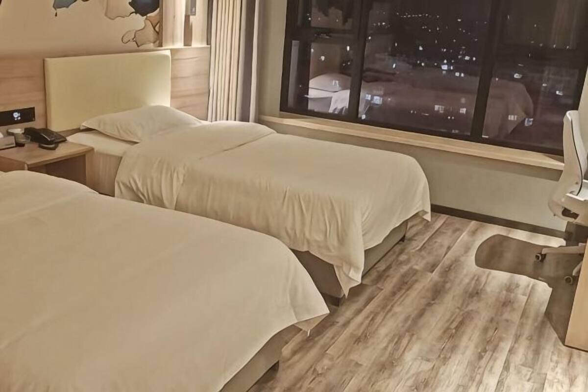 Quarto de hotel com duas camas, mesa de cabeceira com telefone, luminária de mesa e vista para a cidade à noite.