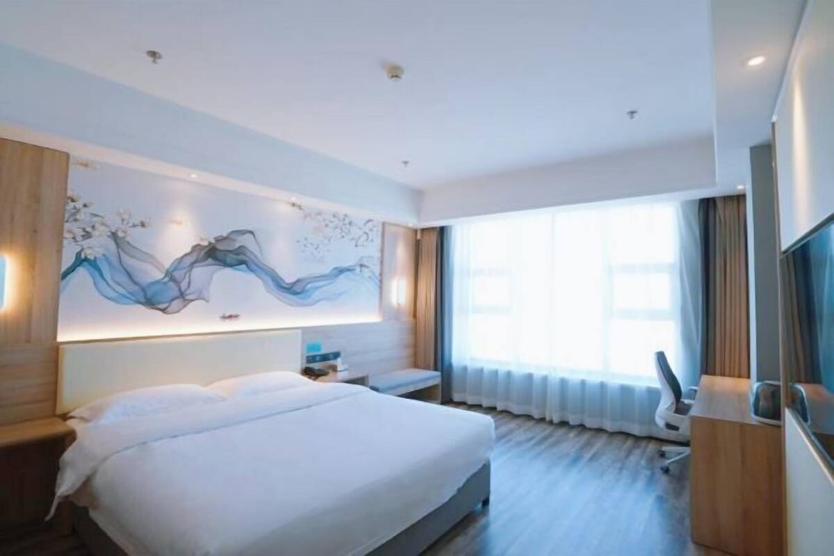 Quarto de hotel moderno com uma cama grande, uma obra de arte pendurada na parede, uma mesinha de cabeceira, uma cadeira e uma janela ampla com cortinas.