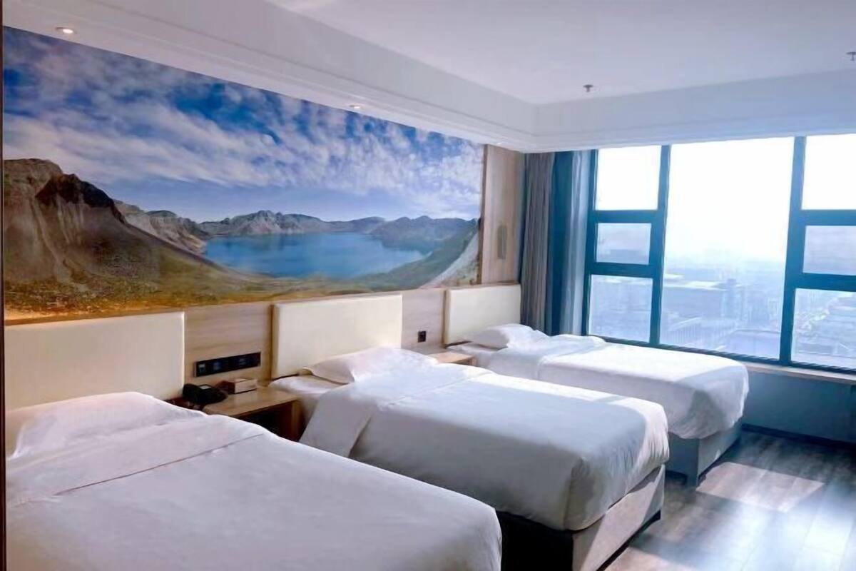 Quarto de hotel com três camas, um grande mural de paisagem montanhosa e uma janela ampla com vista para a cidade.
