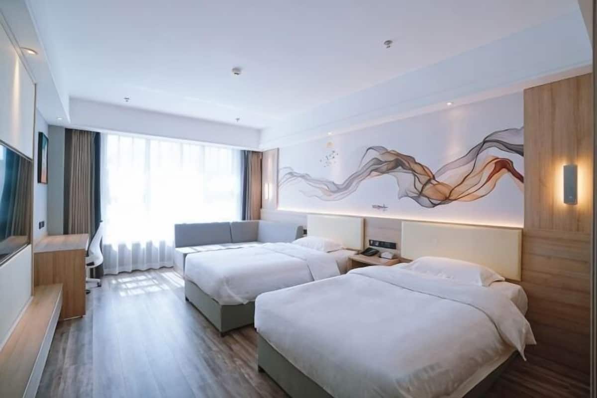 Quarto de hotel com duas camas, uma escrivaninha, uma janela grande com cortinas e uma obra de arte na parede.