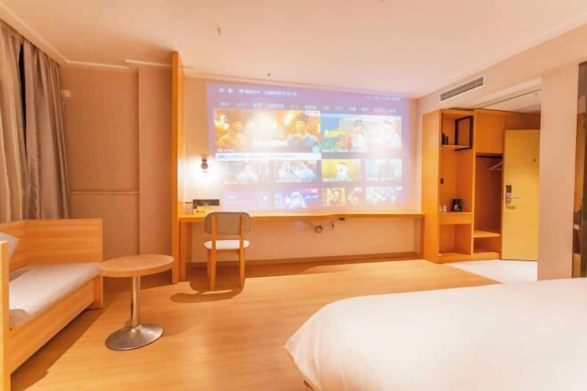 Une chambre d’hôtel comprenant un lit, un bureau avec une chaise, une petite table et un téléviseur fixé au mur diffusant un film.