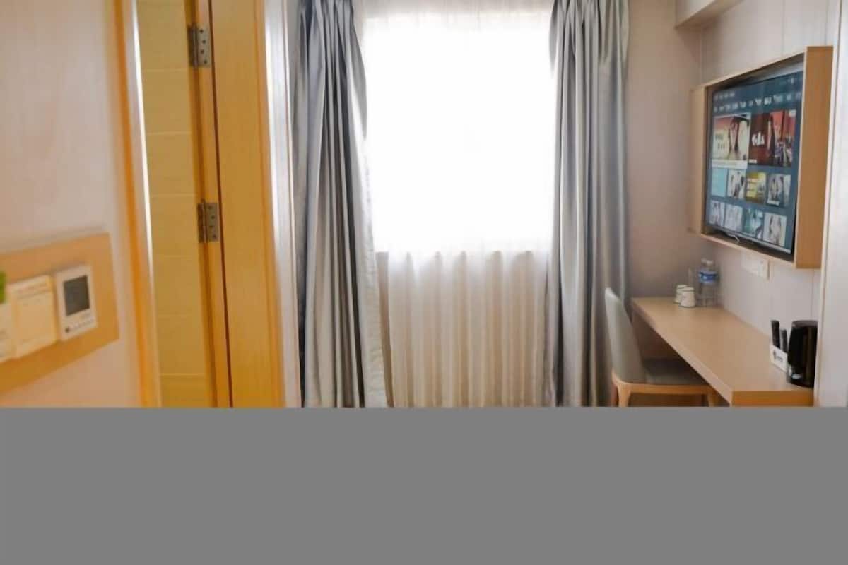 A hotel room with a wooden desk, a small table, and a wall-mounted TV displaying various channels.
