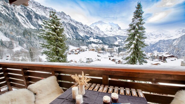 Une terrasse en bois avec une table dressée pour deux personnes, agrémentée de bougies, d’un vase rempli de fleurs séchées et d’une assiette de raisins.