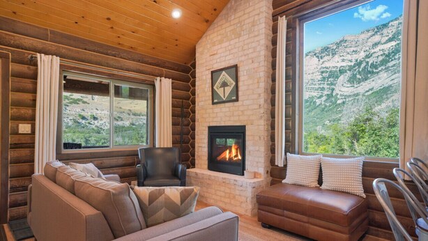 A log cabin interior with a fireplace, a sofa, a chair, and a large window offering a mountain view.
