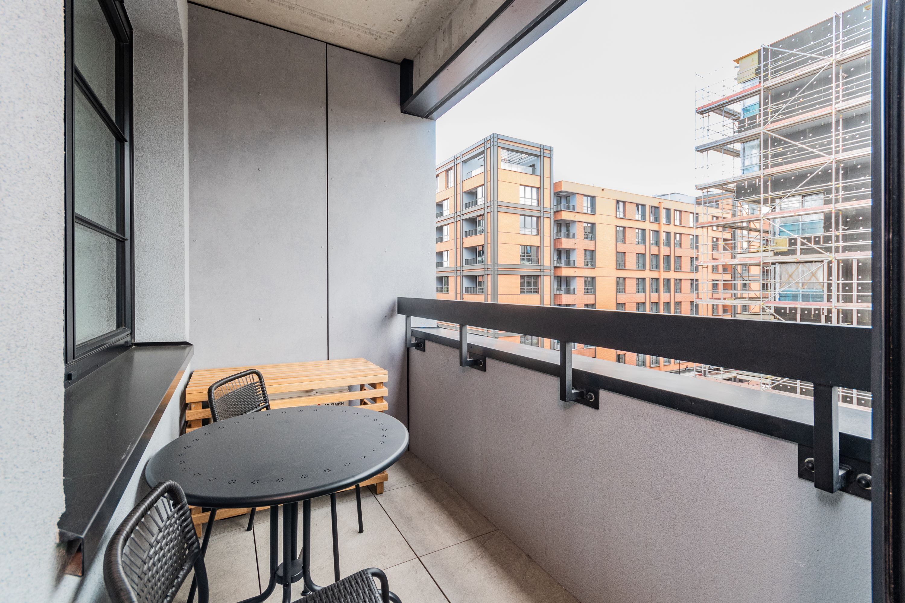 Un balcone con un tavolo rotondo e due sedie, una panca in legno e vista su un edificio a più piani in costruzione.