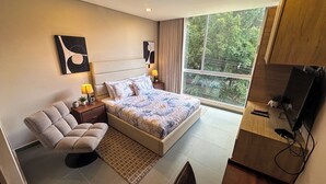 Un dormitorio con una cama grande, una silla, una mesita de noche con lámparas, un televisor y una ventana con vistas a árboles.