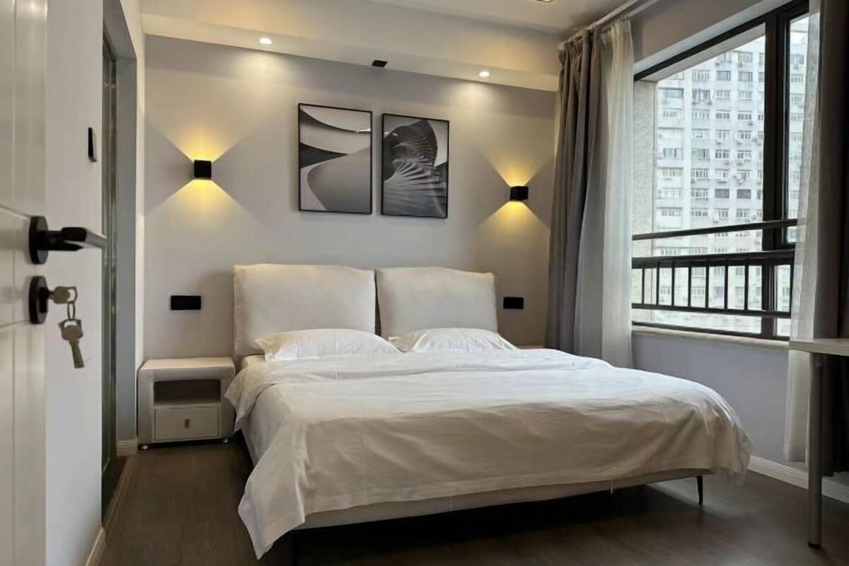 Ein modernes Schlafzimmer mit Bett, Nachttischen, Wandleuchten und einem Fenster mit Blick auf Gebäude.