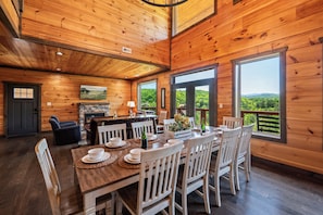 A wooden interior with a dining table set for six, a fireplace, and large windows offering a view of greenery. - Spacious Cabin: Great views, convenient location, theater, game room, hot tub (Sevierville)