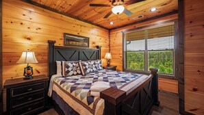Une chambre à coucher dans une cabane en bois, avec un lit, des tables de chevet, un ventilateur de plafond et une fenêtre avec des stores.