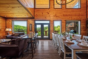 A wooden cabin interior with a dining area, sofa, and large windows.