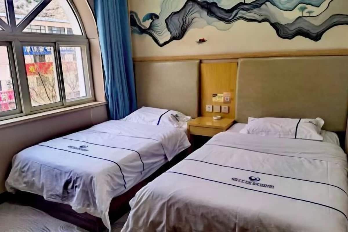 A hotel room with two beds, a window with curtains, a wall mural, and a wooden nightstand with a lamp.