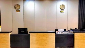 Ένας χώρος υποδοχής με έναν πάγκο υποδοχής, δύο ρολόγια στον τοίχο και μια μαύρη ηλεκτρονική συσκευή πάνω στον πάγκο.