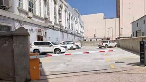 Ένας περιφραγμένος χώρος στάθμευσης με πολλά λευκά αυτοκίνητα παρκαρισμένα, ένα κτίριο με τοξωτά παράθυρα κ αι ένας καθαρός ουρανός.