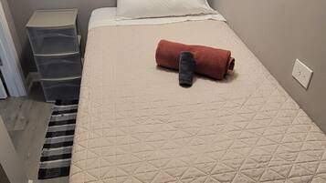 Uma cama de solteiro com colchão acolchoado, uma toalha enrolada e um travesseiro branco. Há um guarda-roupa branco acima da cama e um conjunto de caixas organizadoras cinzas à esquerda.