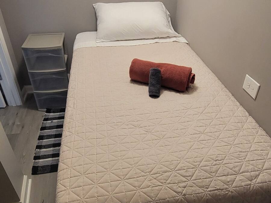 Uma cama de solteiro com colchão acolchoado, uma toalha enrolada e um travesseiro branco. Há um guarda-roupa branco acima da cama e um conjunto de caixas organizadoras cinzas à esquerda.