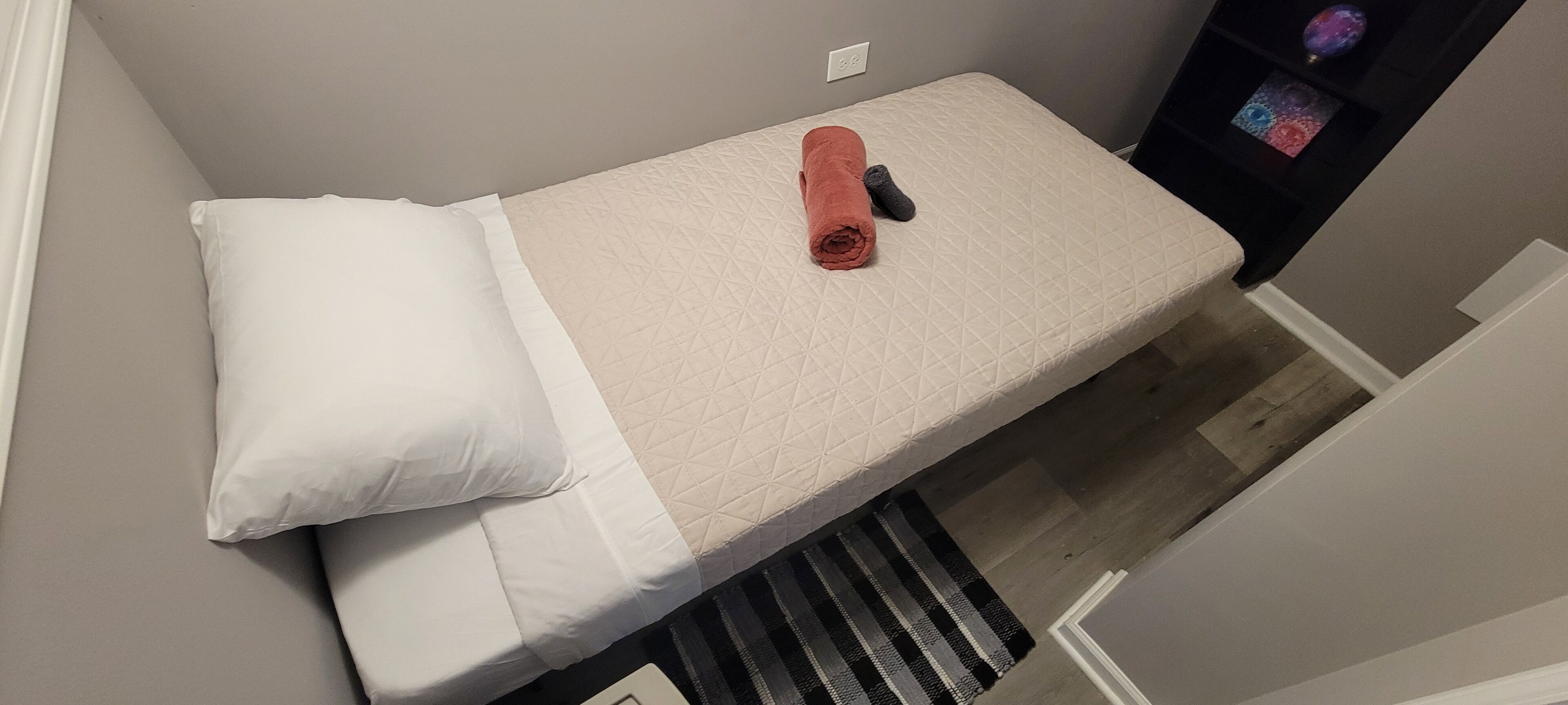 Una cama pequeña y ordenada con un colcha blanca y beige, una toalla enrollada y un objeto negro sobre la cama.