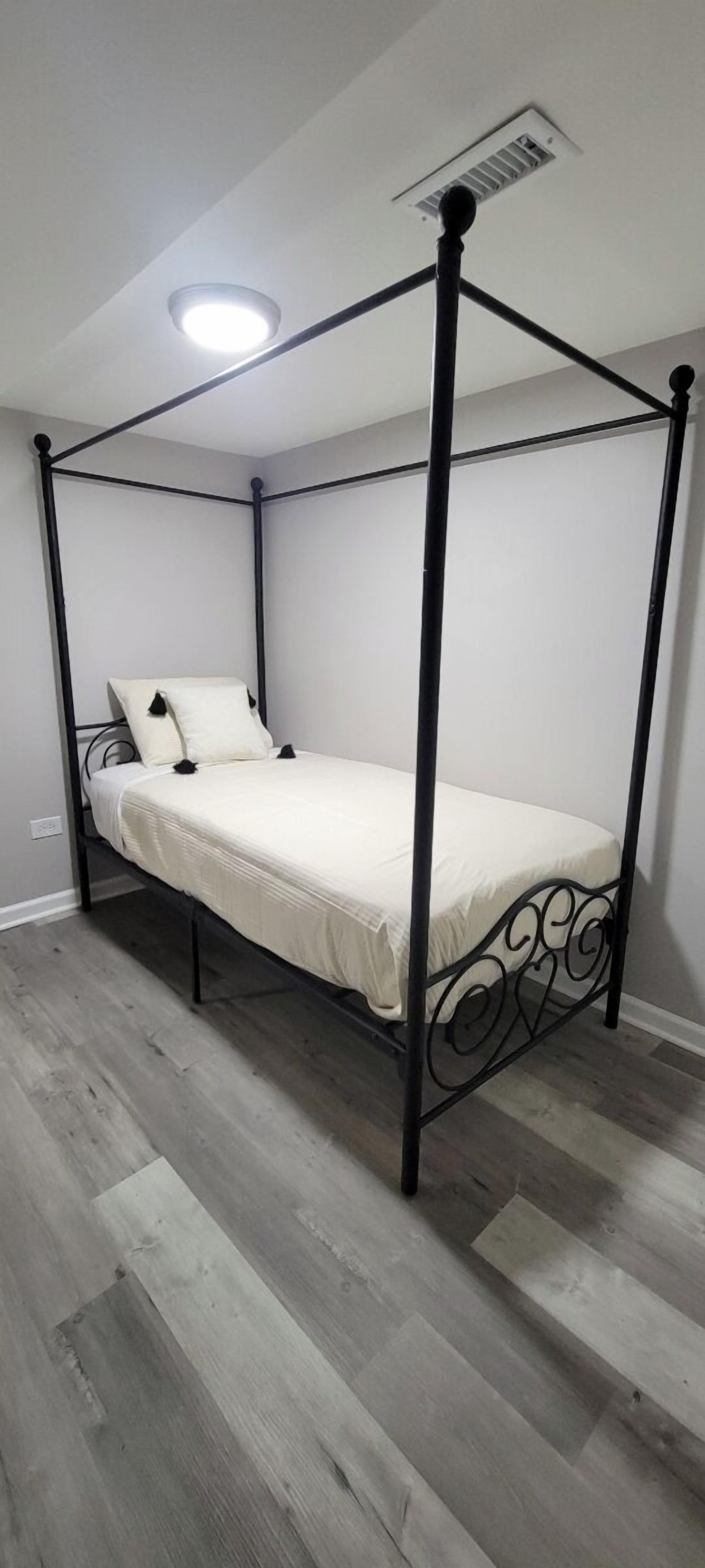 A four-poster bed with a white mattress and pillows, set on a wooden floor in a room with white walls and a ceiling light.