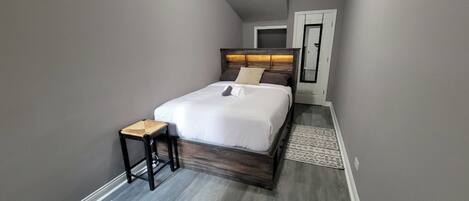 A narrow, modern bedroom with a bed, a small stool, and a door.
