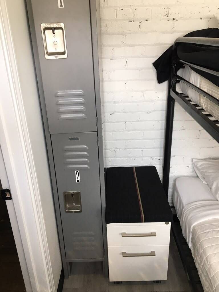 A bunk bed with a black mattress and white bedding, a gray metal locker with a number 2, and a white drawer unit.